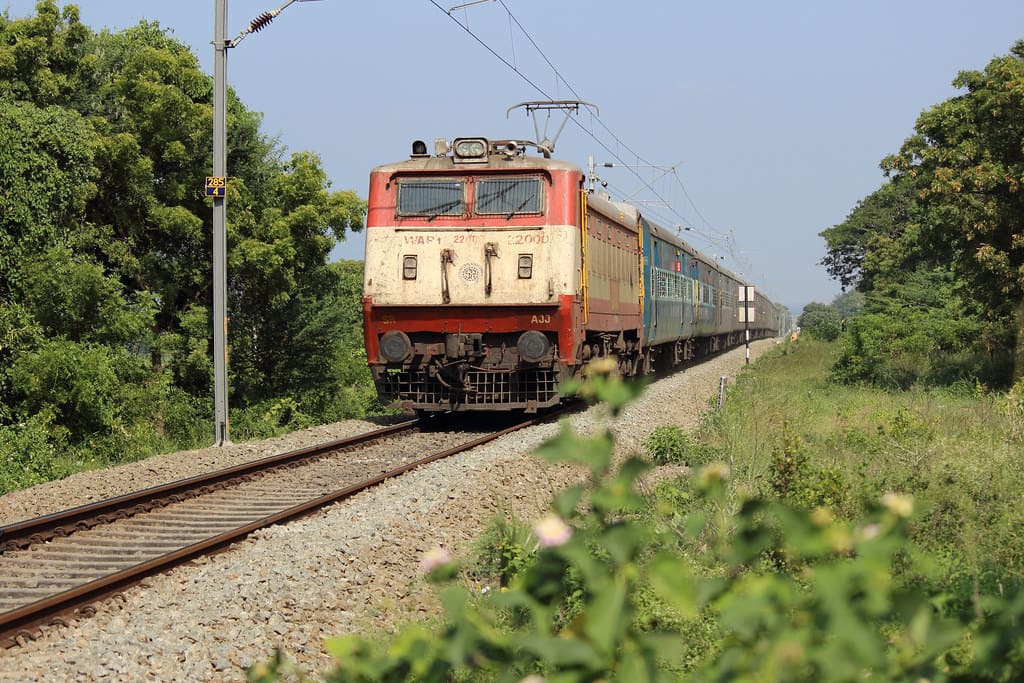 Indian Railways passenger train running on single track line through rural landscape with electric locomotive