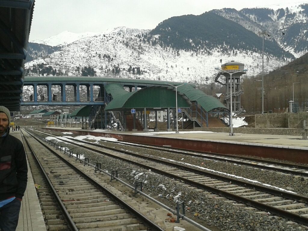 Snow-covered railway station in Kashmir with mountain backdrop and train tracks in winter conditions