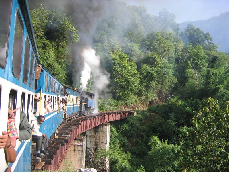 Train crossing a narrow curved bridge through dense forest hills in India with passengers looking out