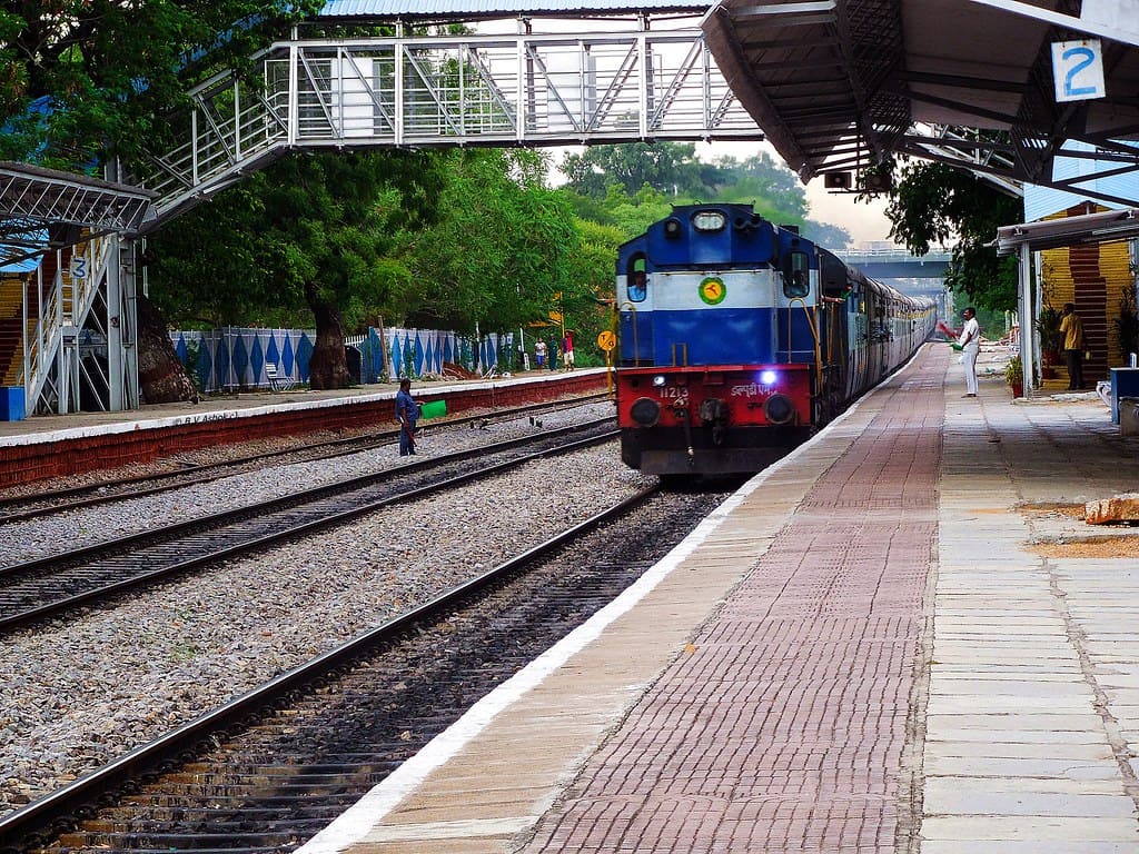 Indian Railways diesel locomotive approaching station platform with passenger train and foot overbridge