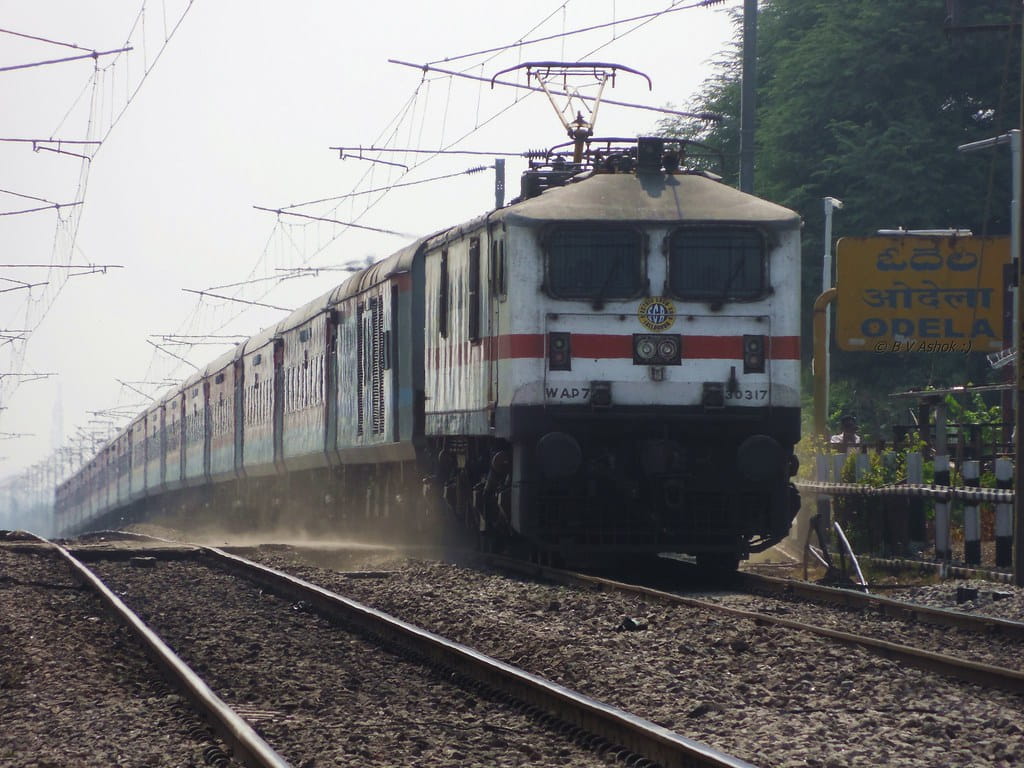 Indian Railways electric locomotive pulling a long passenger train on open track near station