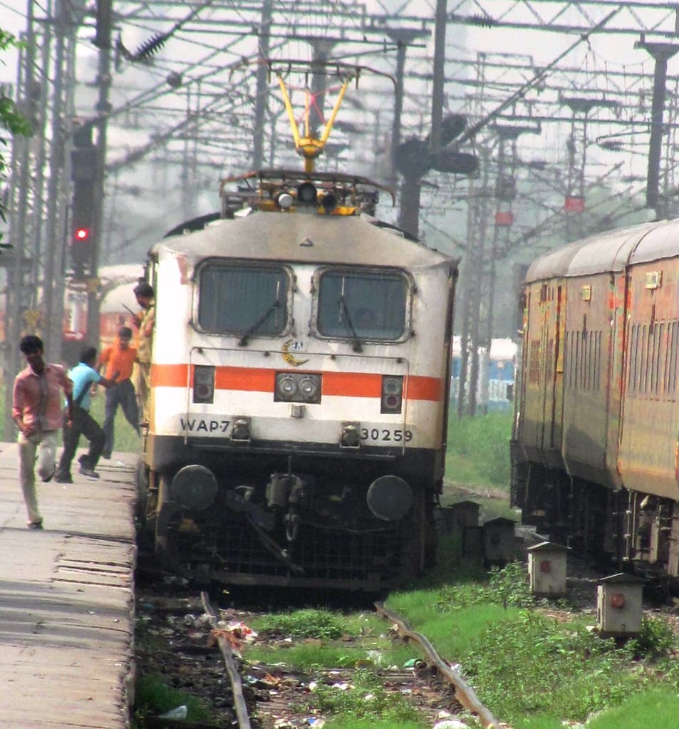 Indian Railways WAP-7 electric locomotive approaching platform with passenger train and overhead electric lines
