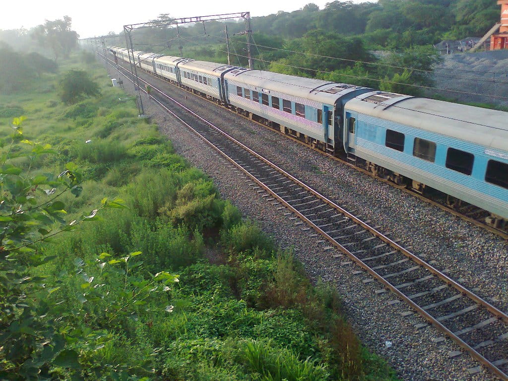 Indian Railways passenger train passing through green countryside with overhead electric lines
